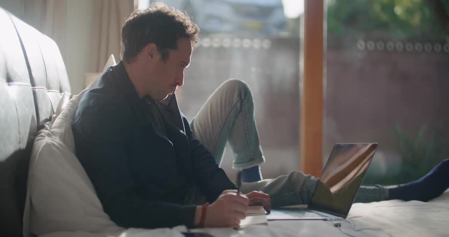 A man is sitting in bed while writing notes as he looks at his laptop screen