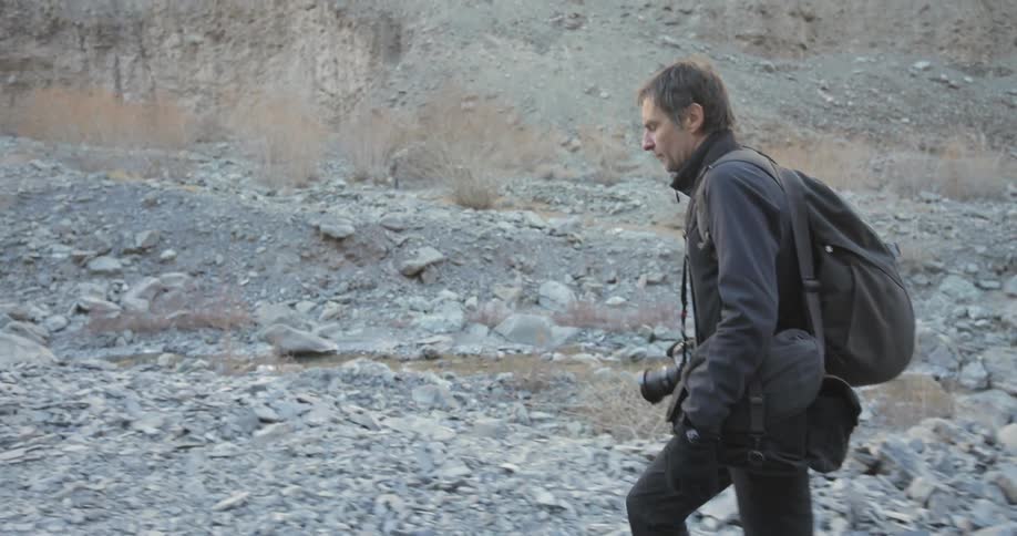 A man with camera equipment walks over a rocky path on an overcast day