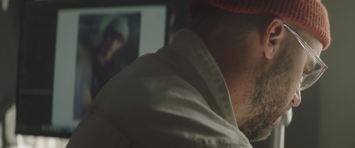 A man with a hat and glasses in a room with a computer screen in the background