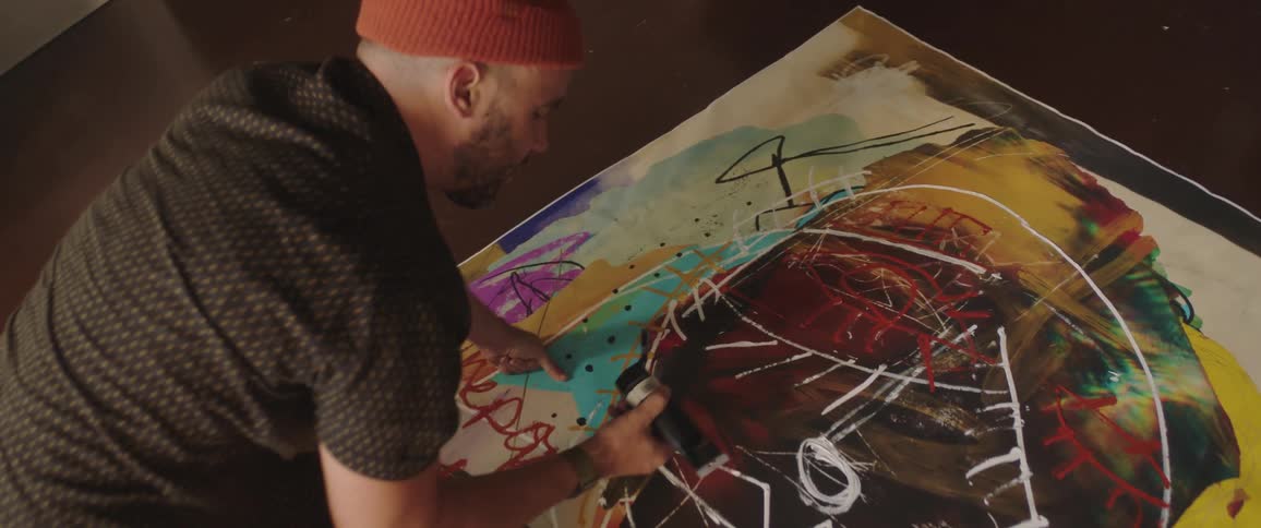 An artist kneeling on the floor adds paint from a tube to his artwork