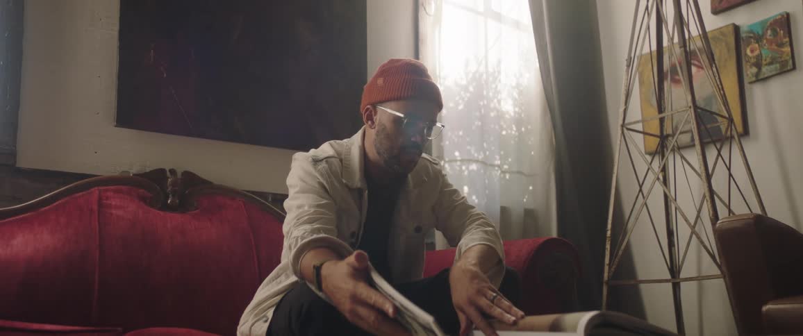 A man with a hat in a room sitting on a couch looking through a book with a wall and a window in the background
