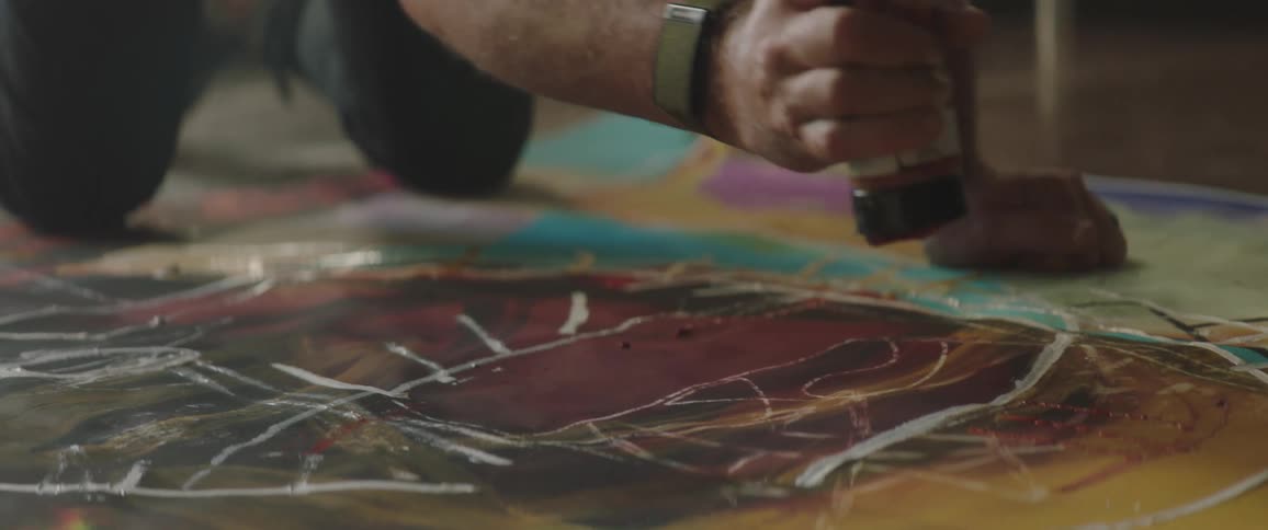 Man wearing a toque kneels on the floor and applies paint from a tube to his artwork