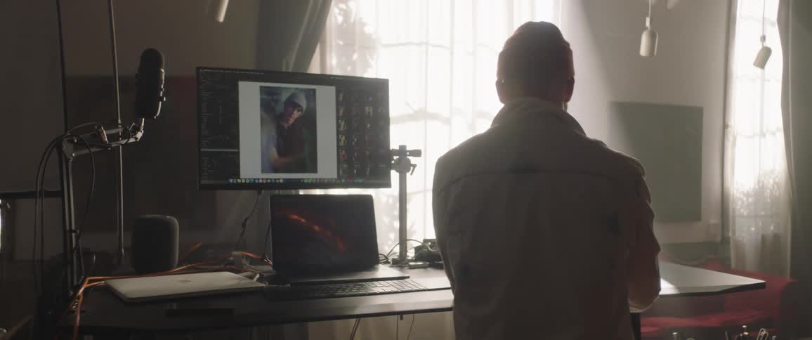 A man with a hat in a room sitting at a desk with computer screens and a microphone