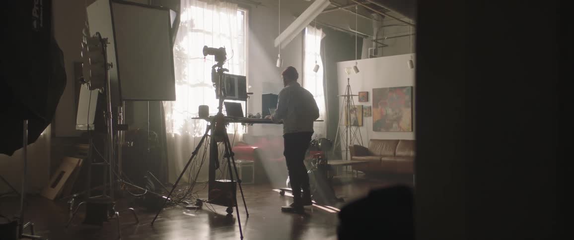A man walks over to a table in a room with photography equipment and starts working on a laptop during the day