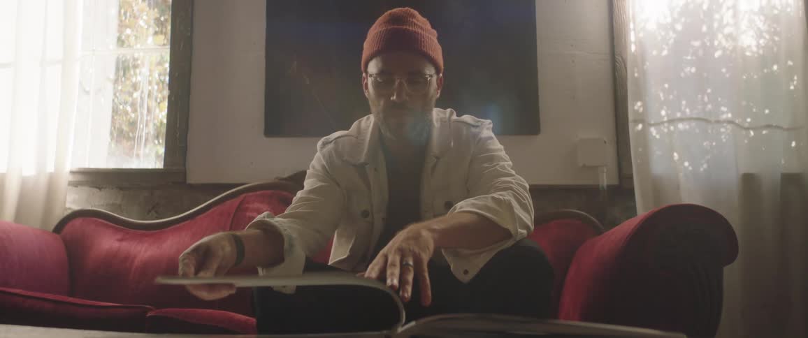 Man wearing a toque and eyeglasses sits on a red sofa and turns the pages of an album