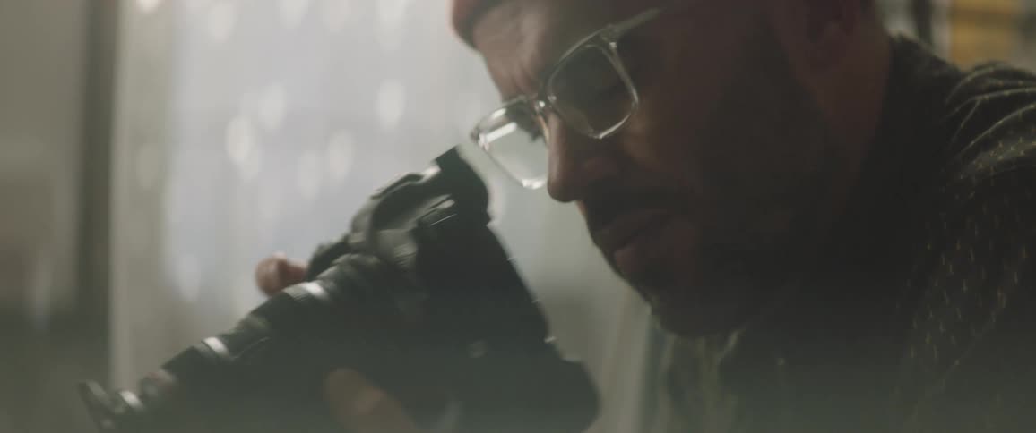 A man with a hat and glasses in a room taking a picture with a camera