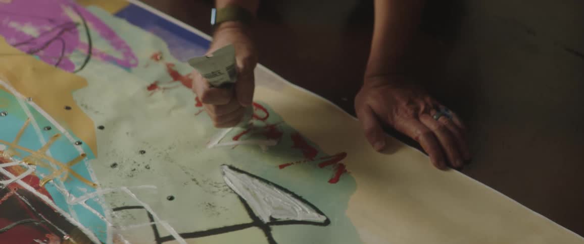 Man applies paint from a tube onto an art canvas while kneeling on the floor