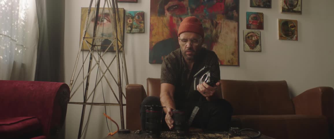 A man in a room sitting on a couch attaching a camera lens to a camera