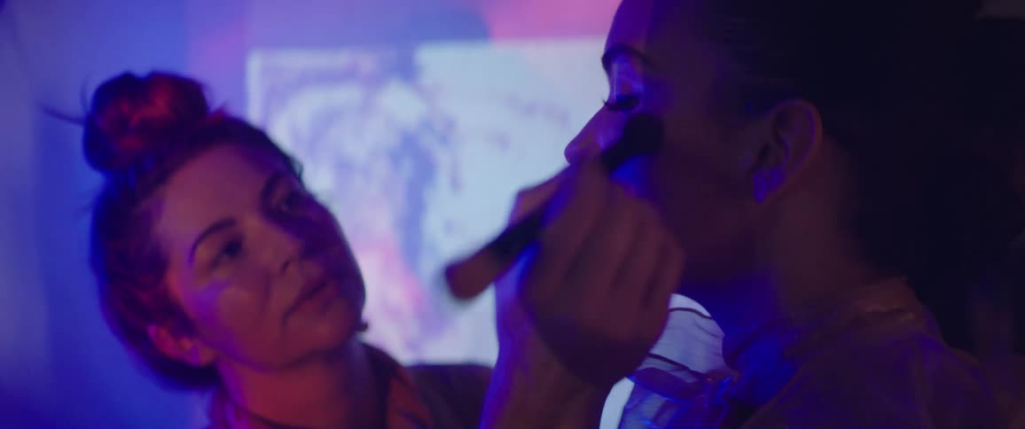 A woman applies makeup with a small brush to another womans face inside of a woman with a flashing projector screen