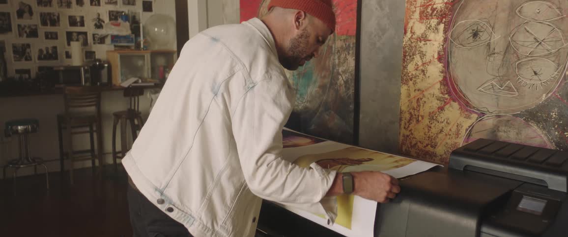 A male artist looks closely at a large poster sized print of an artistic photograph atop the poster printer