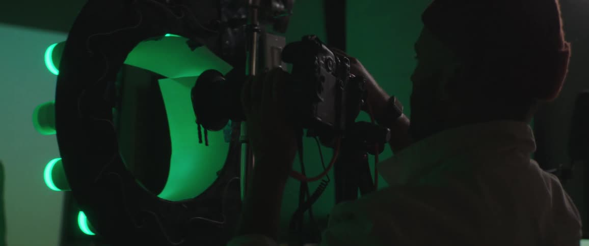 A photography stands behind a lighting device and positions a camera taking a photo of a male model in a photoshoot room