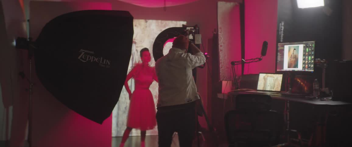 A woman poses in front of a backdrop with changing light colors as a man photographs her