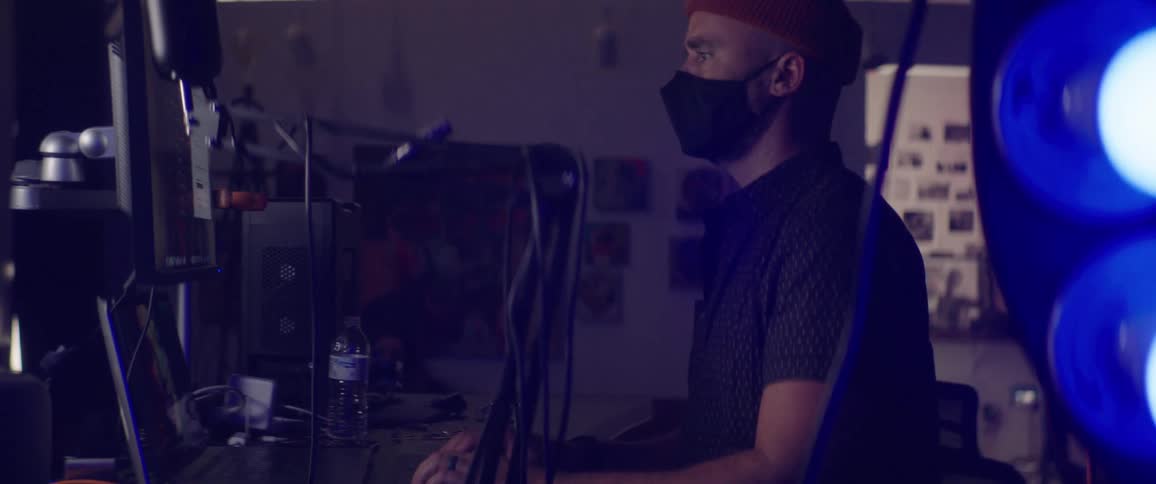 A man in a medical mask sits before a computer at desk covered in computer equipment