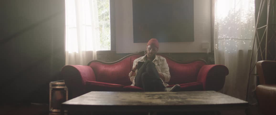 A man with a hat in a room sitting on a couch with a wall and windows in the background