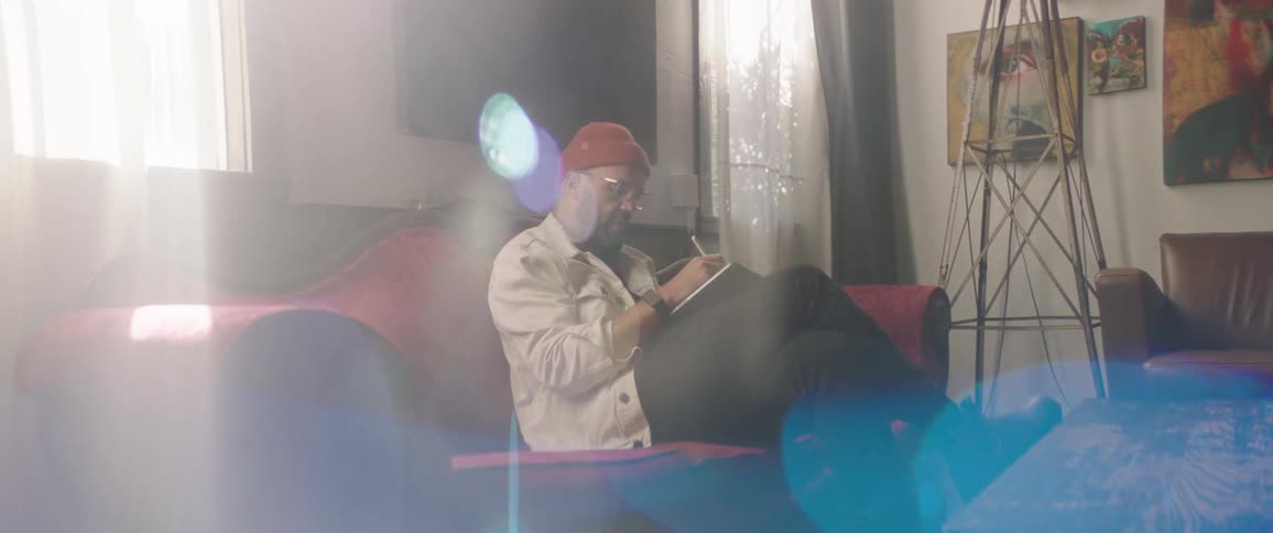 Man sits on sofa between windows drawing on tablet as sun rays shine through windows