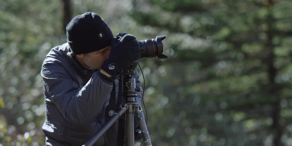 A man crouches by a camera on a tripod