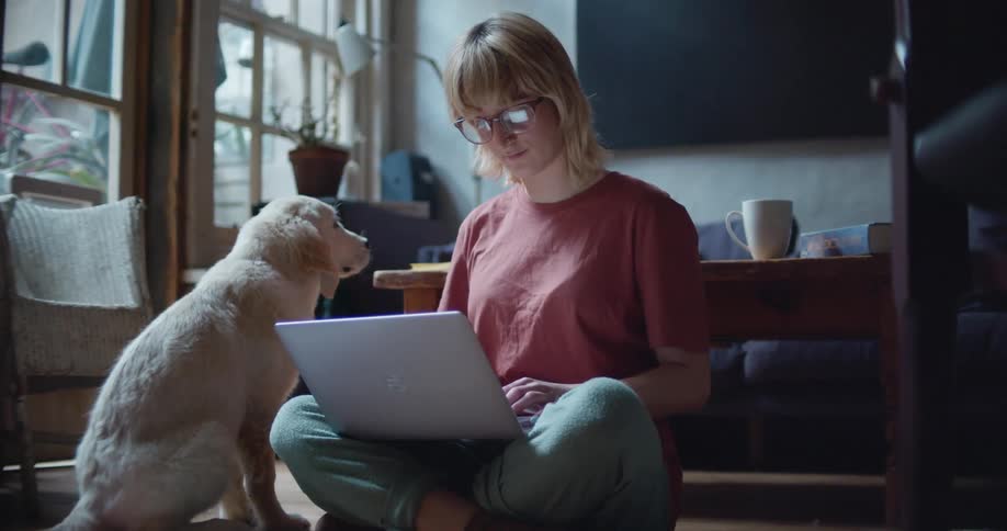 A woman with glasses sitting on the floor using a laptop and petting a puppy