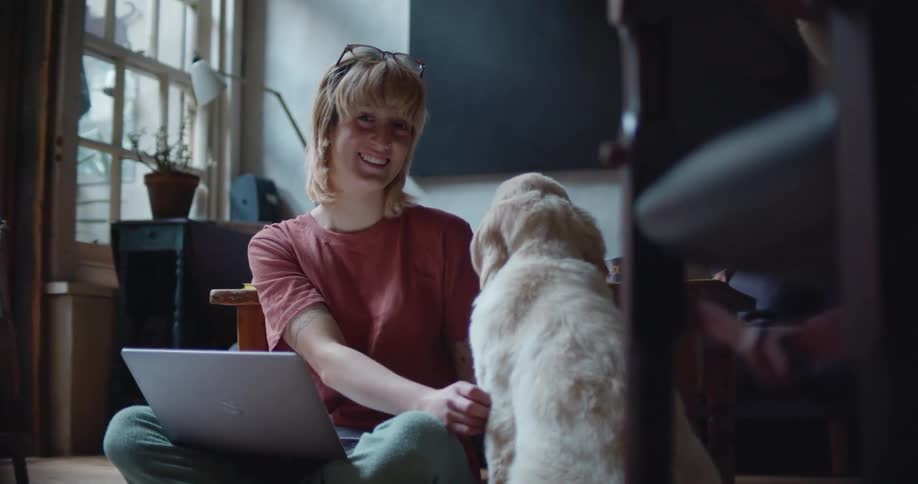 Blonde woman sits on floor with laptop in her lap petting dog smiling and laughing