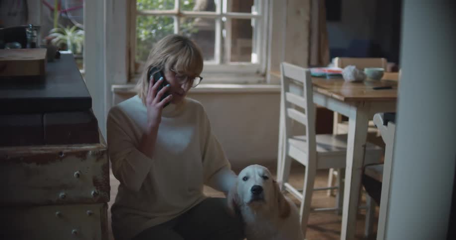 A woman with glasses in a room talking on a smart phone while petting a dog