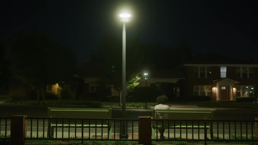 A lone female sits on a bench next to a street at night and a car passes by