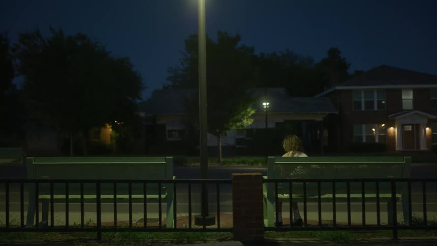 Woman sits waiting on bench near street across from large homes at night