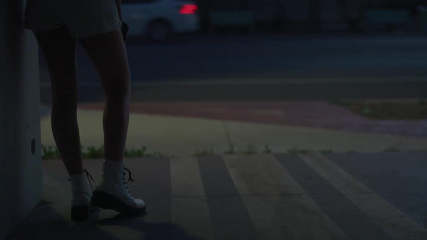 Woman wearing shorts leans against building at night as traffic passes along nearby street