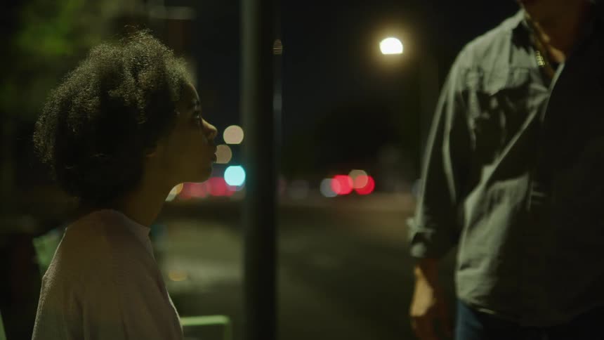 Man walking parallel to street stops to talk to woman sitting on bench at night