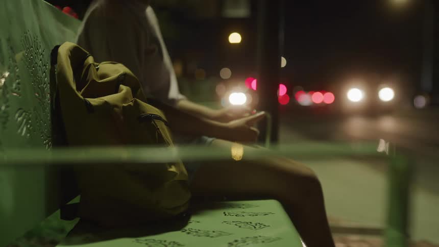 Woman with bag sits on bench at side of road as traffic passes at night