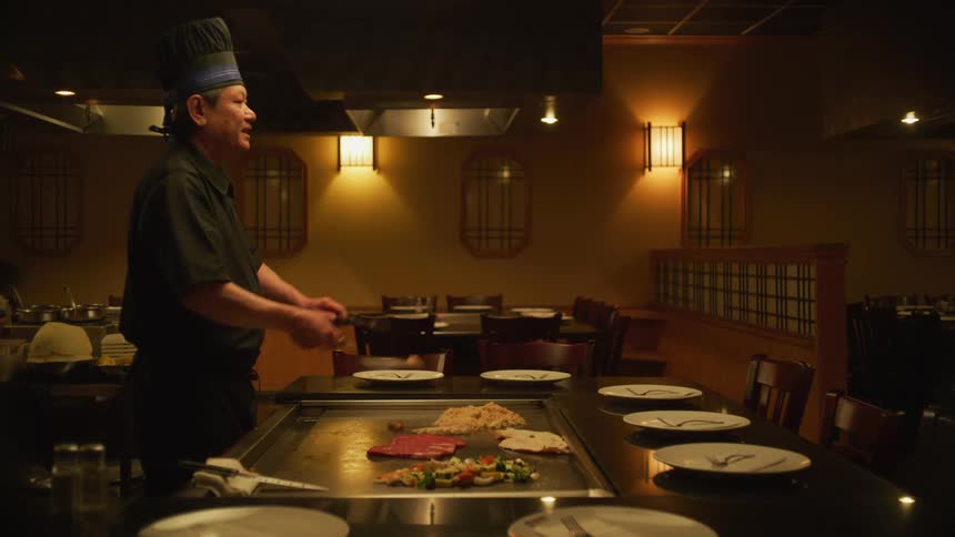 Chef stands in front of grill full of food before throwing a piece of meat into air in practice motion but no one is in empty restaurant to catch it