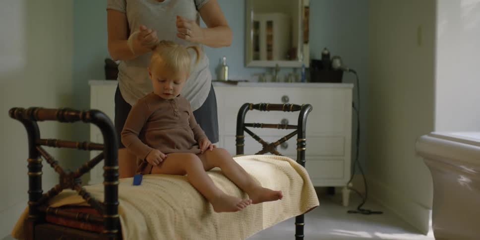 A woman in a bathroom styling a little girls hair with a wall and mirror in the background