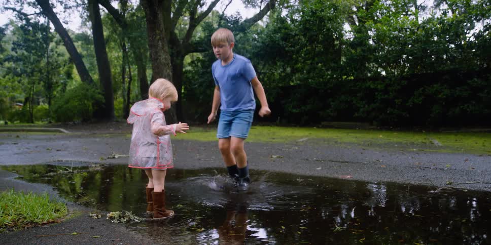 A boy and a small girl playfully jump in a puddle in a road outdoors during the day