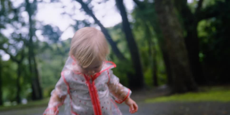 A blonde toddler splashing in an outdoor puddle while wearing a rain jacket and boots