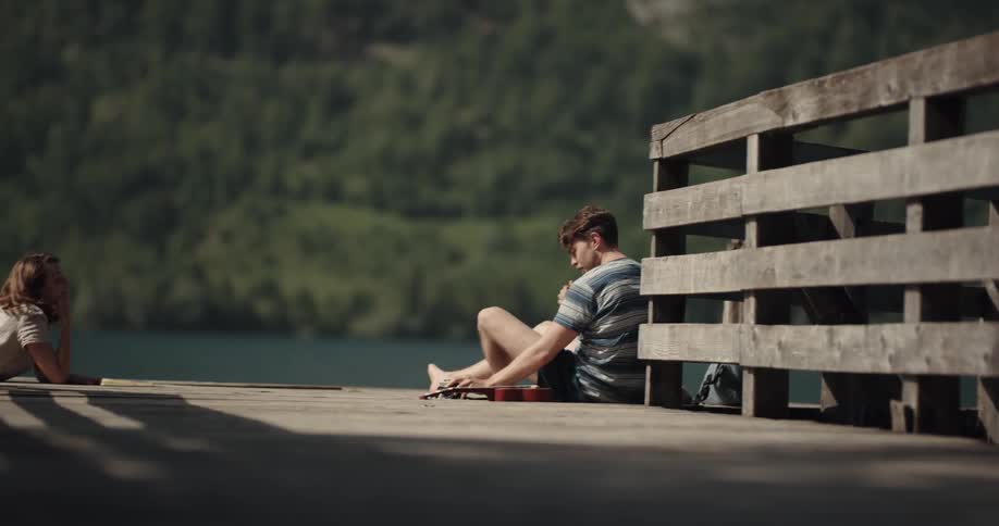 A man and a woman on a dock with the man playing a ukulele and taking a picture with a camera