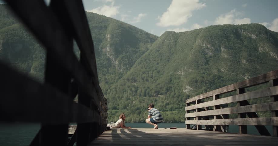 A man taking a picture of a woman lying down at the end of a dock outdoors next to a lake a mountain side