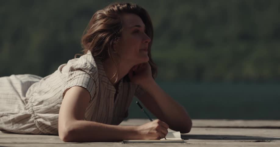 A young woman rest on her stomach on a wooden dock while writing in a notepad