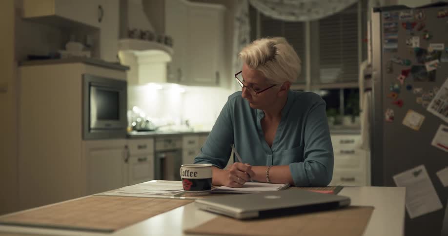 A woman sits at a kitchen table seemingly stressed out while writing on a piece of paper