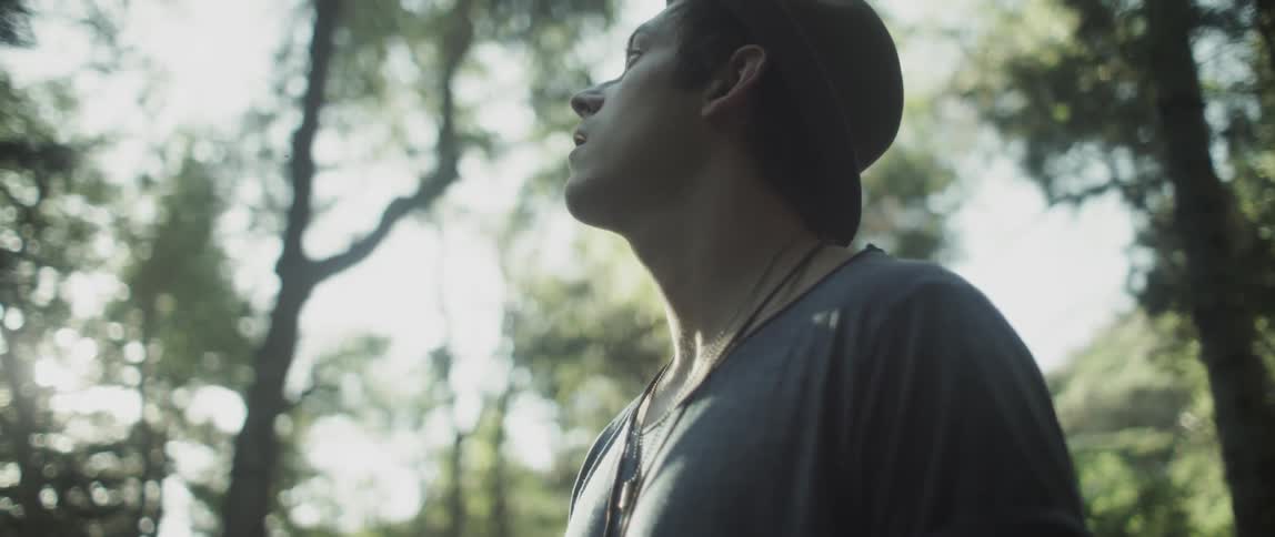 A man wearing a hat is standing in a wooded area with sun blazing through the trees