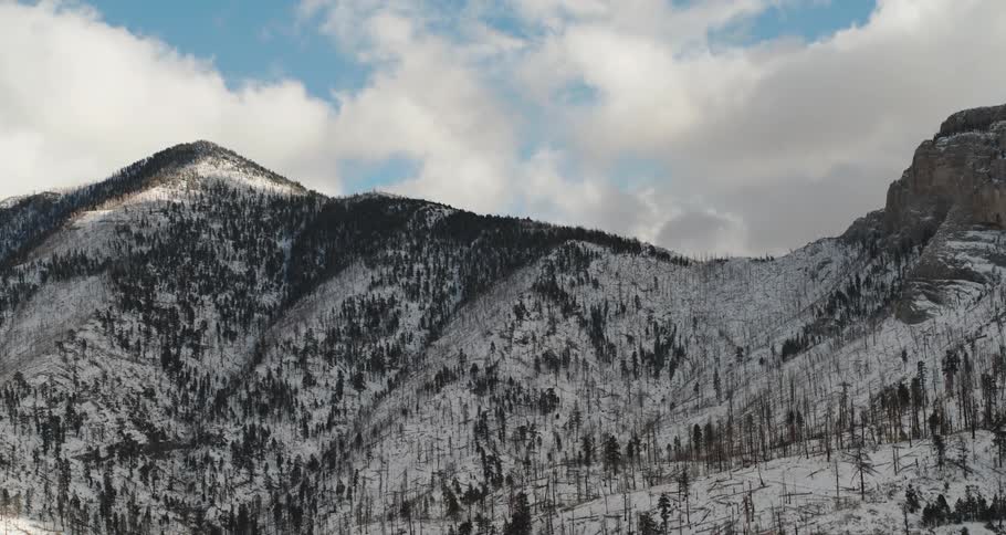 A mountain ridge dusted with snow and trees under a bright blue sky and white clouds