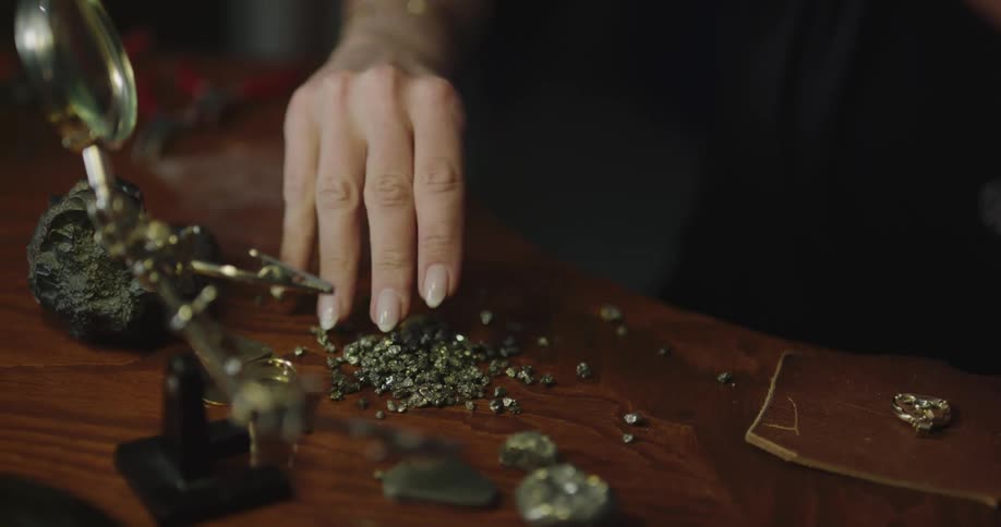 A woman drops gem like stones on a table then picks up tweezers to help add them to a ring