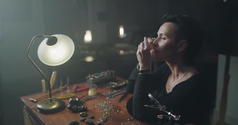 A woman uses a jewelers glass to examine a stone while sitting at a table with tools and gems