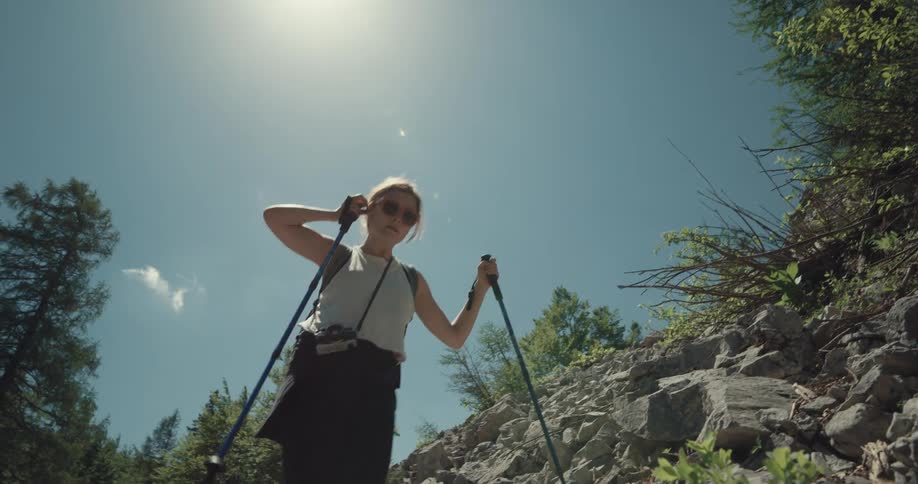 A woman walking on the side of a rocky hill with two trekking poles in a forest during the day