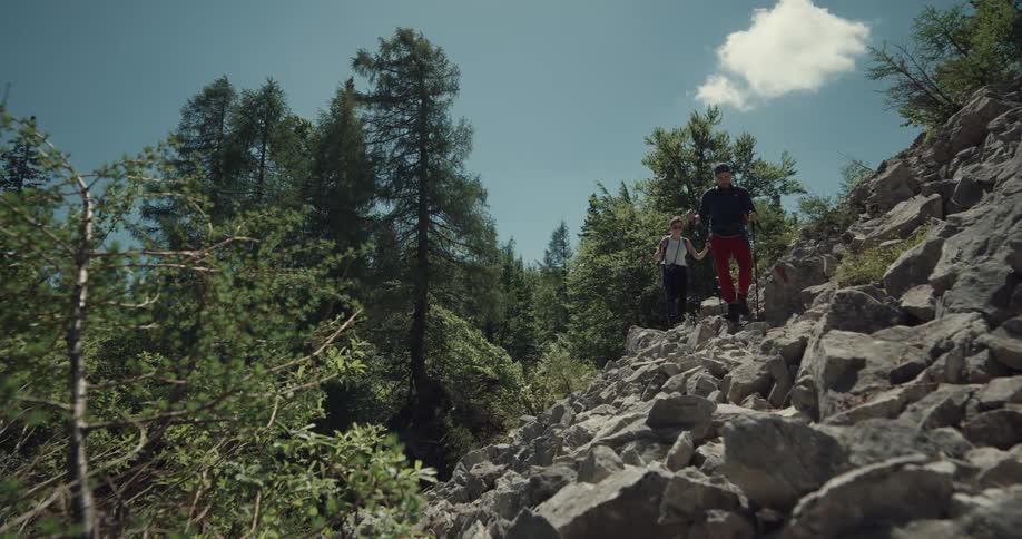 Man and woman hike along rock strewn steep slope near conifer trees under slightly cloudy sky