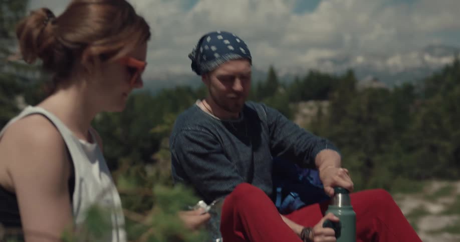 A man and a woman sitting down outdoors in the wilderness of a forest next to a hill packing away their hiking gear during the day