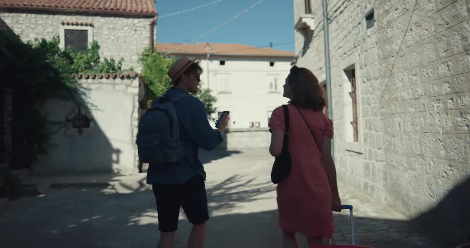 A man and woman walk through a town street with travel bags looking around like tourists