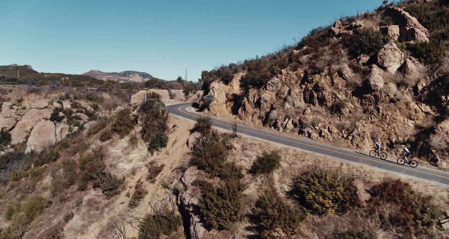 Two people are riding bicycles on a winding mountain road with mountains in the distance