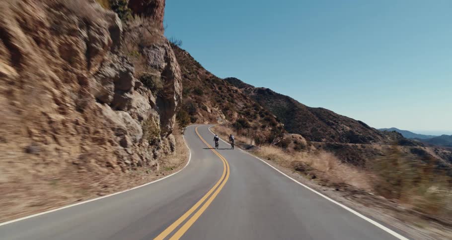 Two people riding bicycles on the highway near a cliff with the sky and clouds in the background