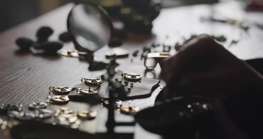 A woman examines a gem stone under a magnifying glass then places it in jewelry with tweezers