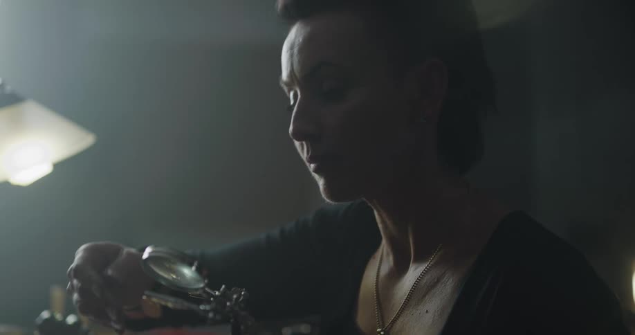 A woman with a necklace in a room using a magnifying glass to look at a jewelry piece with a lamp in the background