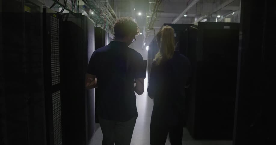 A man and woman walk through a server room holding a tablet talking and laughing