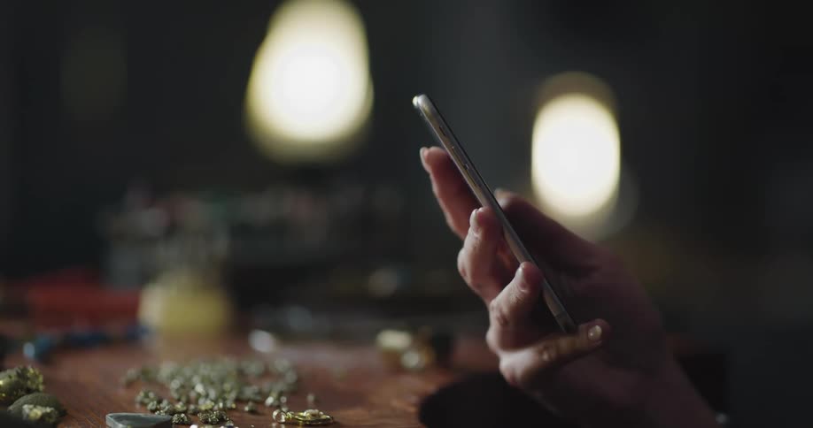 A woman picks up her smartphone while sitting at a table covered with beads